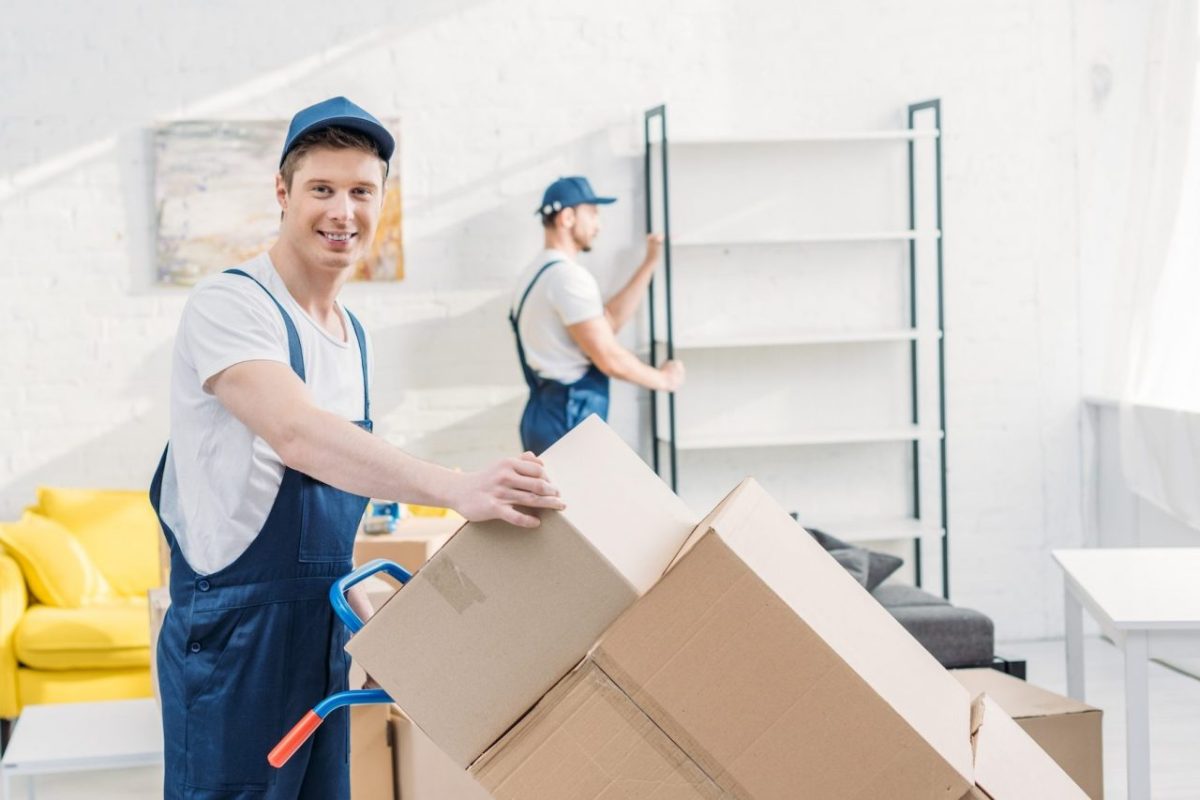 two-movers-in-uniform-transporting-cardboard-boxes-and-furniture-in-apartment-e1670221445352.jpg